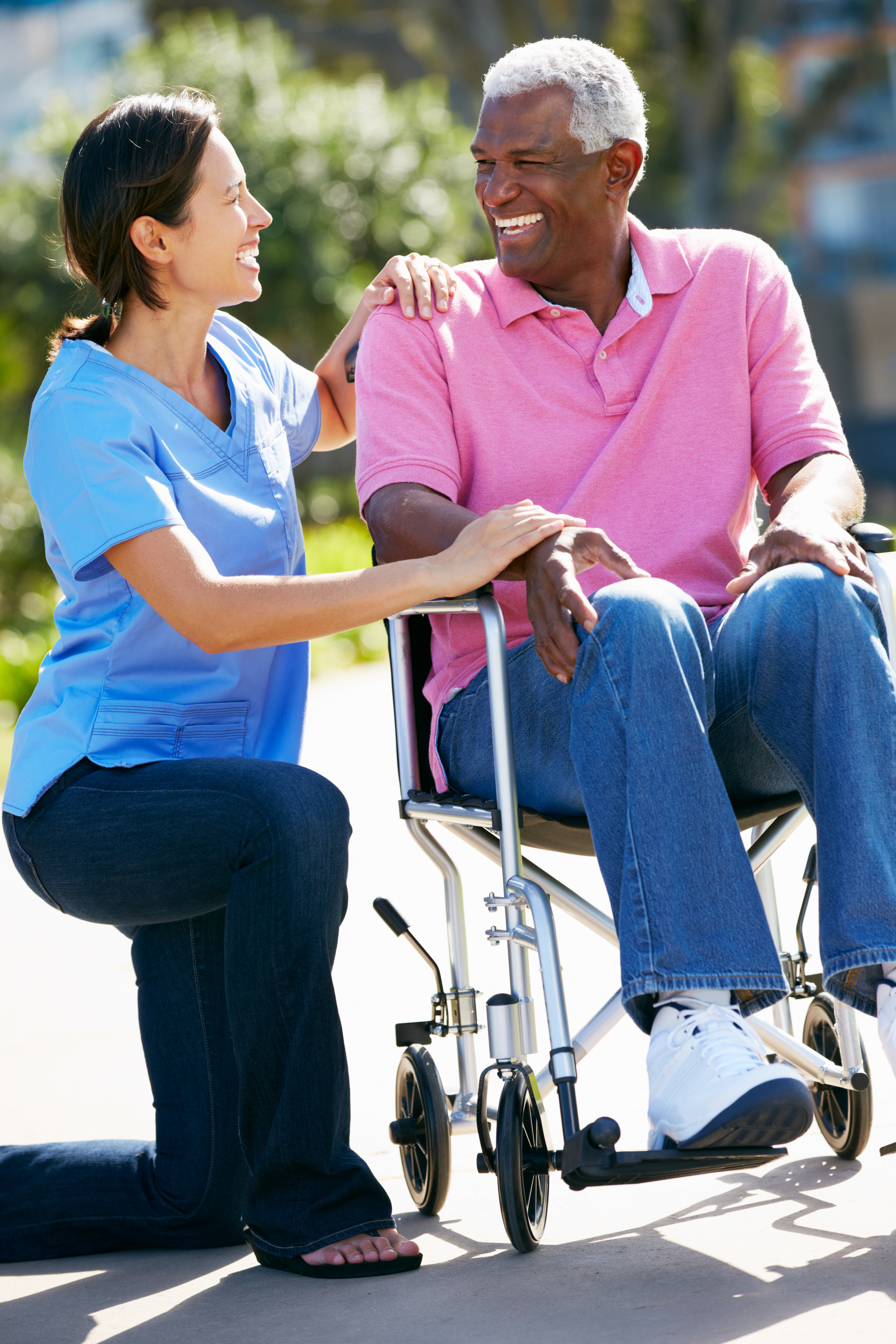 A caregiver in blue scrubs kneels beside an elderly man