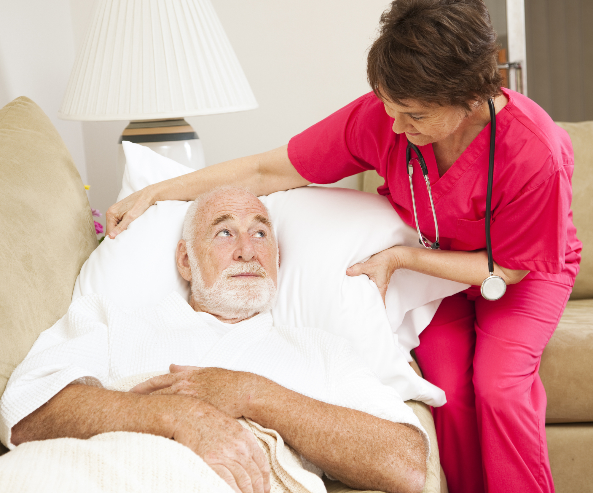 Elderly man lying on a couch with a white robe is tended to by a nurse